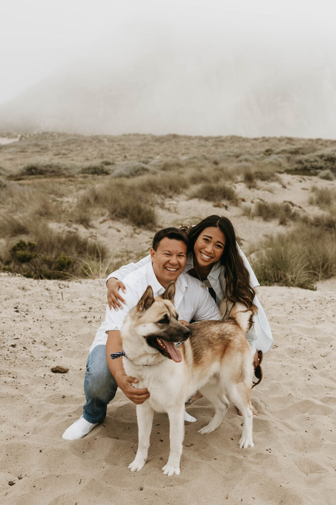 couple posing with their dog on a beach for their moody engagement photoshoot

